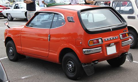 Rear three-quarter of an orange small hatchback with a black-framed rear window, visitors behind.