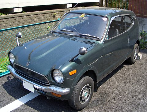 Dark green small coupé from the front left, round headlamps, parked by a wire fence.