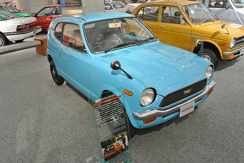 Light blue small coupé in a museum hall with an information panel by its front wheel, a yellow Z behind. Opens a larger view