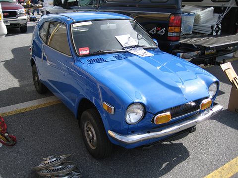Blue small coupé with a for-sale sign in the windscreen, stalls and parts around it. Opens a larger view