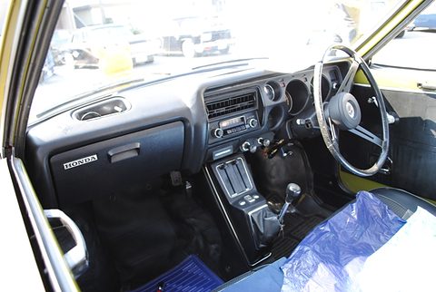 Inside a small car: a three-spoke wheel, round dials, a black centre console and a plastic-covered seat.