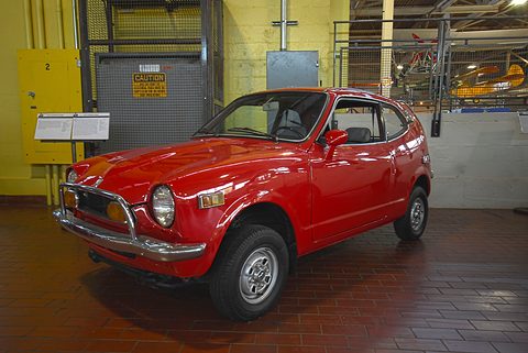 Red small coupé on a brown tiled floor in front of a yellow wall and a roller door. Opens a larger view