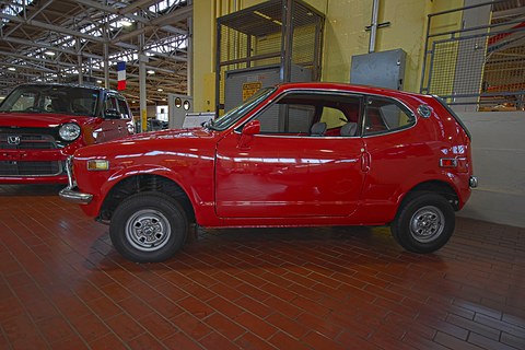 Side view of a red small coupé on a brown tiled floor, other cars behind. Opens a larger view
