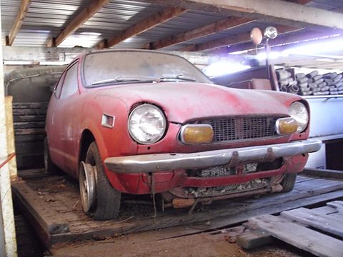 A dusty faded red small coupé on a flatbed under a corrugated roof. Opens a larger view