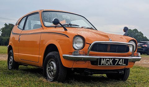 Orange small coupé with black side stripes and a black-and-silver number plate, on grass. Opens a larger view