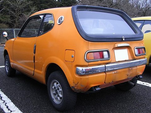 Rear three-quarter of an orange hatchback with a black-framed rear window, bare trees behind. Opens a larger view