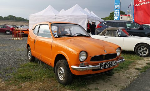 Orange small coupé parked on grass and gravel in front of white marquees. Opens a larger view