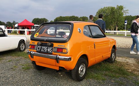 Rear three-quarter of an orange hatchback on gravel, visitors walking past behind. Opens a larger view
