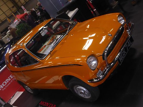 Orange small coupé with black side stripes on a club stand in a dark hall. Opens a larger view