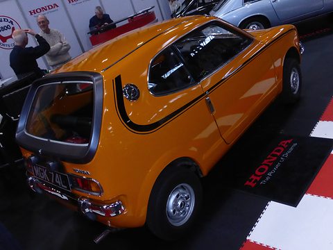 Rear three-quarter of an orange hatchback with black stripes on a chequered stand floor. Opens a larger view