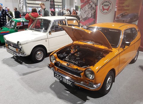 An orange small coupé with its bonnet raised next to a white small saloon on a show stand. Opens a larger view