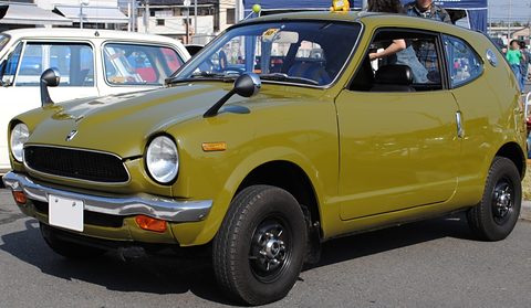 Olive small coupé with round headlamps and a black grille, a white car parked behind. Opens a larger view