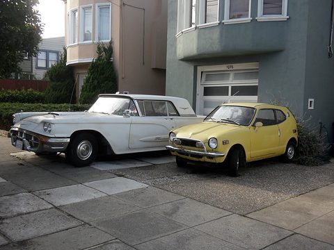 A yellow small coupé parked on a steep driveway in front of a garage, a white Thunderbird beside it. Opens a larger view