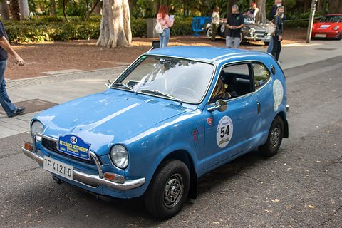 Blue small coupé with a rally plate and number 54 on the door, parked on a street with spectators. Opens a larger view
