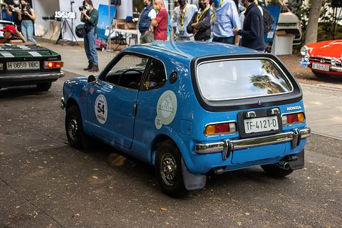 Rear view of a blue hatchback with a rally roundel, spectators and a stand behind. Opens a larger view