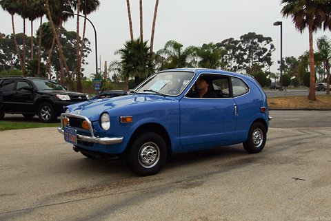 Blue small coupé moving across a car park, palm trees and other cars behind. Opens a larger view