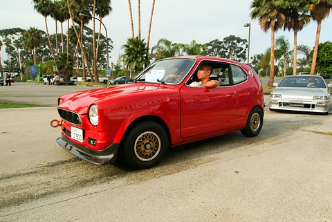 Red small coupé with gold wheels driving across a car park, palm trees behind. Opens a larger view