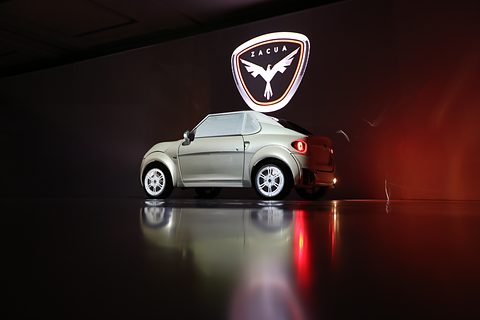 Silver small two-seat car on a dark stage reflecting its lamps, a lit eagle logo above it.
