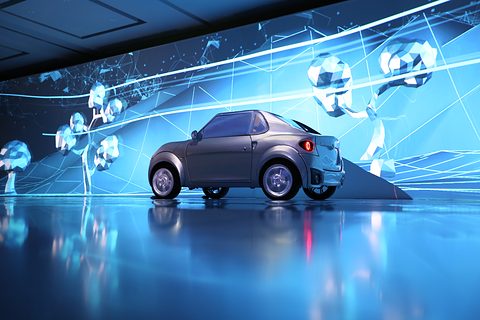 Grey small two-seat electric car on a glossy stage before a huge blue projection of low-polygon trees. Opens a larger view