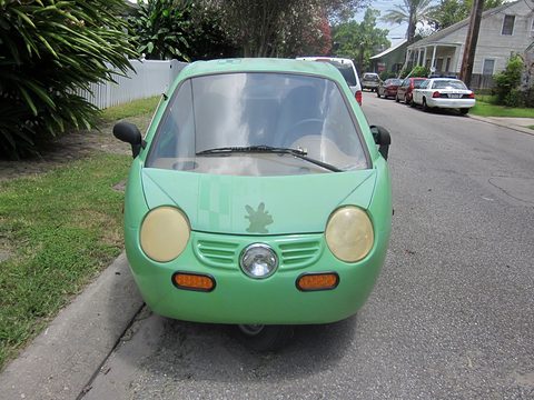 Front of a pale green rounded microcar with large round headlamps and one central front wheel at a kerb.