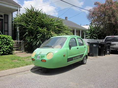 Pale green three-wheeled four-door microcar with round headlamps and ELECTRIC lettering at a kerb by palms. Opens a larger view
