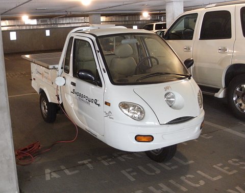 White three-wheeled microcar with a small pickup bed and a charging cable on a car-park floor painted with lettering. Opens a larger view