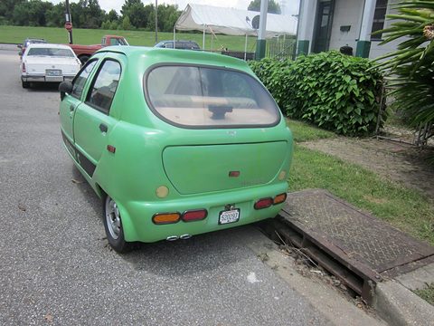 Rear of a pale green rounded microcar with a tailgate and small rear lamps parked beside a drain. Opens a larger view