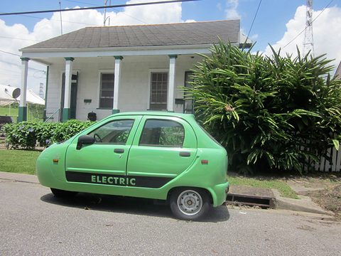 Side view of a pale green four-door microcar with a black ELECTRIC stripe, a white house behind. Opens a larger view