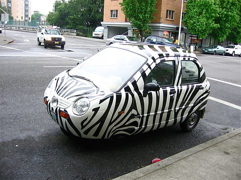 White microcar covered in black zebra stripes parked at a kerb on a wet street, trees and buildings behind.