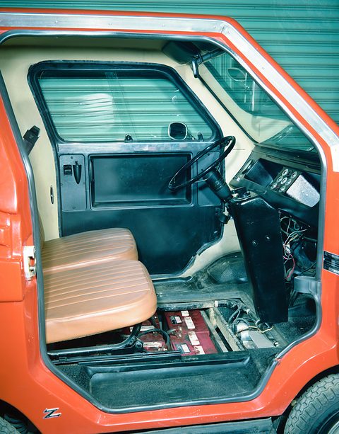 Open door of an orange car revealing tan pleated seats, a black dashboard and a row of batteries sunk into the floor.