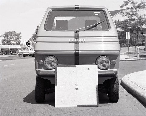 Black-and-white front view of a square small car with a plywood board leaning on its bumper, a road and trees behind. Opens a larger view