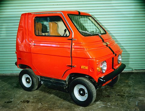 Orange boxy two-seater with black bonnet stripes, a black bumper and round headlamps, parked in front of a green roller door. Opens a larger view