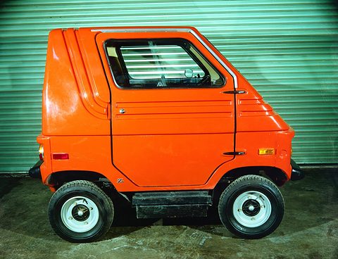 Orange tall square two-seater in exact side profile in front of a green roller door, white wheels. Opens a larger view
