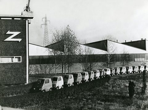 A row of small boxy cars stretching along a factory lawn beside a tall building with a large Z sign.
