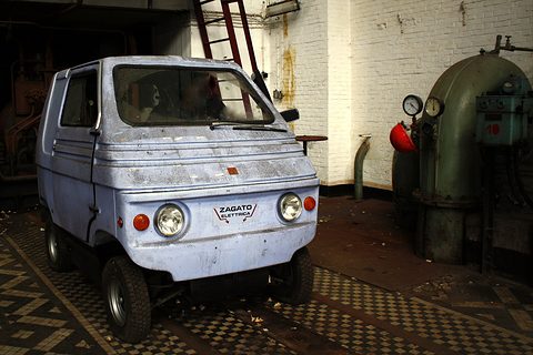 Dusty pale blue boxy two-seater with scuffed paint on a tiled floor beside an old green pump and a white brick wall. Opens a larger view
