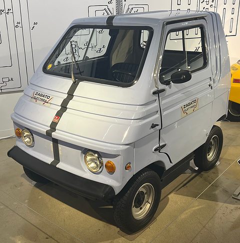 Pale grey boxy two-seater with a black stripe, round headlamps and Zagato Elettrica door badges, in front of a white wall printed with diagrams.