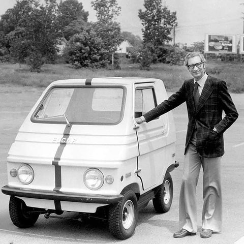 A man in a check jacket with his hand on the door of a small white boxy car with grey stripes over its nose.