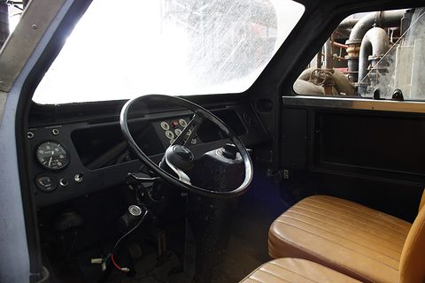 Dusty black dashboard with round gauges, a large black steering wheel and tan pleated seats behind a scratched windscreen. Opens a larger view