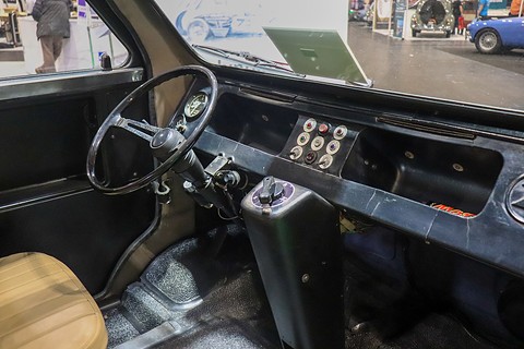 Black dashboard with a thin-rimmed steering wheel, a small switch panel and a selector lever on a central tunnel, beige seat at left.