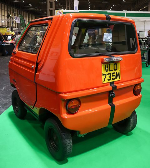 Rear three-quarter view of a tall orange box-shaped car with a large rear window, round tail lamps and a black stripe. Opens a larger view