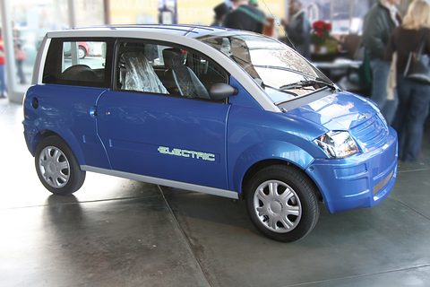 Bright blue small hatchback with a silver roof frame and ELECTRIC lettering on a showroom floor.