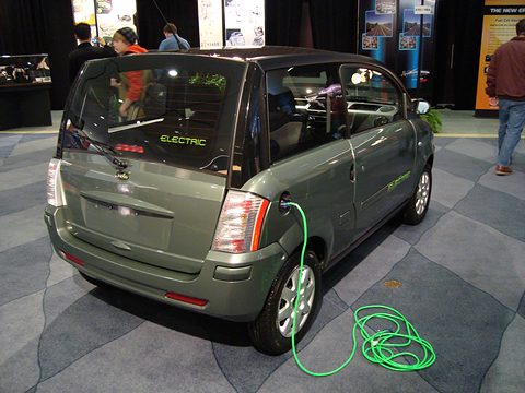 Rear of a grey-green small hatchback with a bright green charging cable coiled on a patterned carpet. Opens a larger view