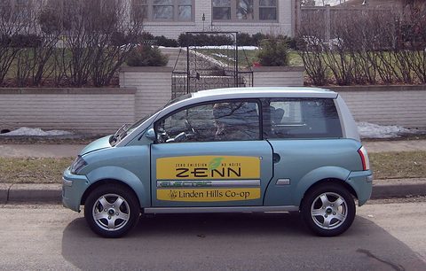 Side view of a light-blue small hatchback with a ZENN door panel parked by a wall and an iron gate. Opens a larger view