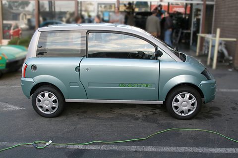 Side view of a pale blue-grey small hatchback with ELECTRIC lettering and a green charging cable on the road. Opens a larger view
