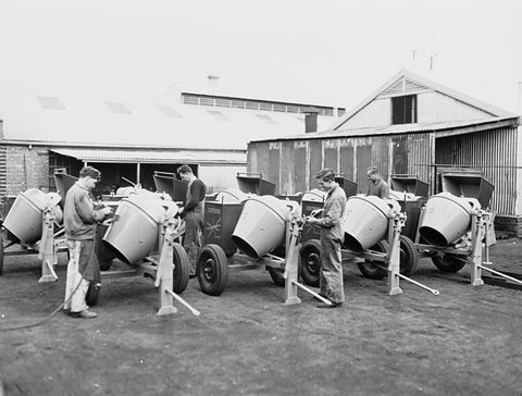 Black-and-white photograph of four men in overalls tending a row of new concrete mixers outside corrugated-iron sheds.