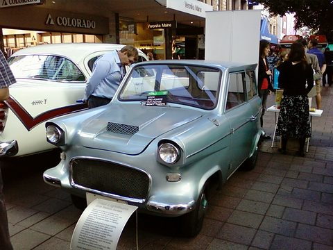 Pale blue small saloon parked on brick paving beside a white car, a man leaning over it and shoppers behind. Opens a larger view