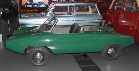Side view of a long low green roadster with no doors, a pale blue saloon parked behind it. Opens a larger view